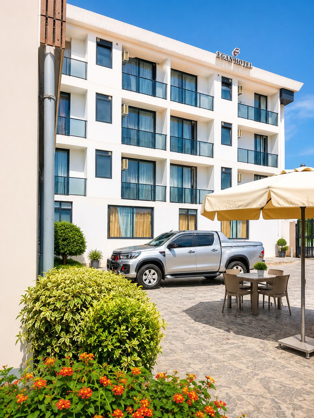 A hotel building with a sign reading 'EGAN HOTEL,' a silver pickup truck parked in front, outdoor seating with a table and chairs, and a well-maintained garden with bushes and flowers.