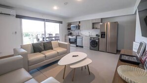 Een modern appartement met een woonkamer, bank, salontafel en keuken inclusief een koelkast, magnetron en wasmachine.
