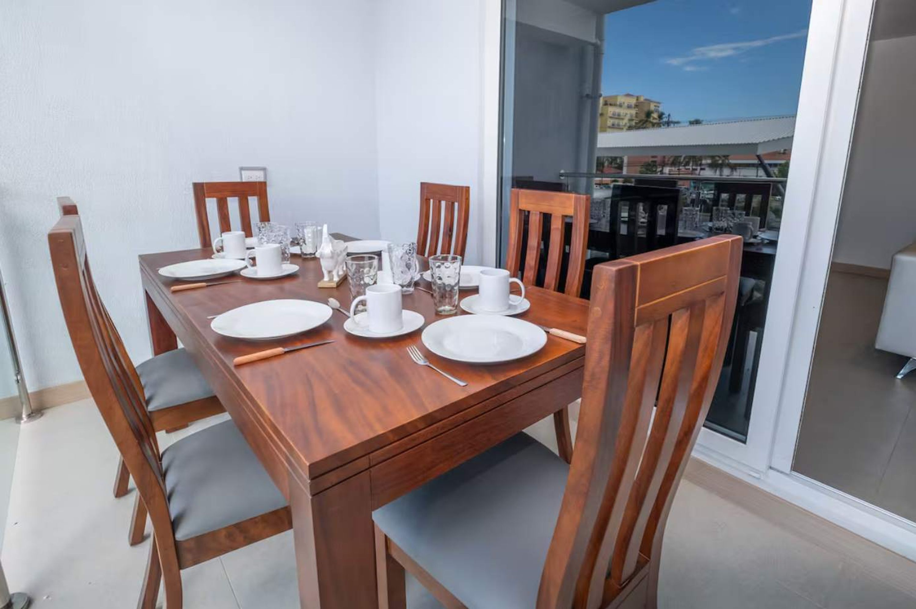 Une table à manger dressée avec de l’assiette blanche, des tasses et des couverts, entourée de chaises en bois avec des coussins gris.