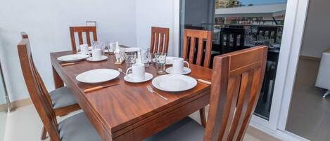 Une table à manger dressée avec de l’assiette blanche, des tasses et des couverts, entourée de chaises en bois avec des coussins gris.