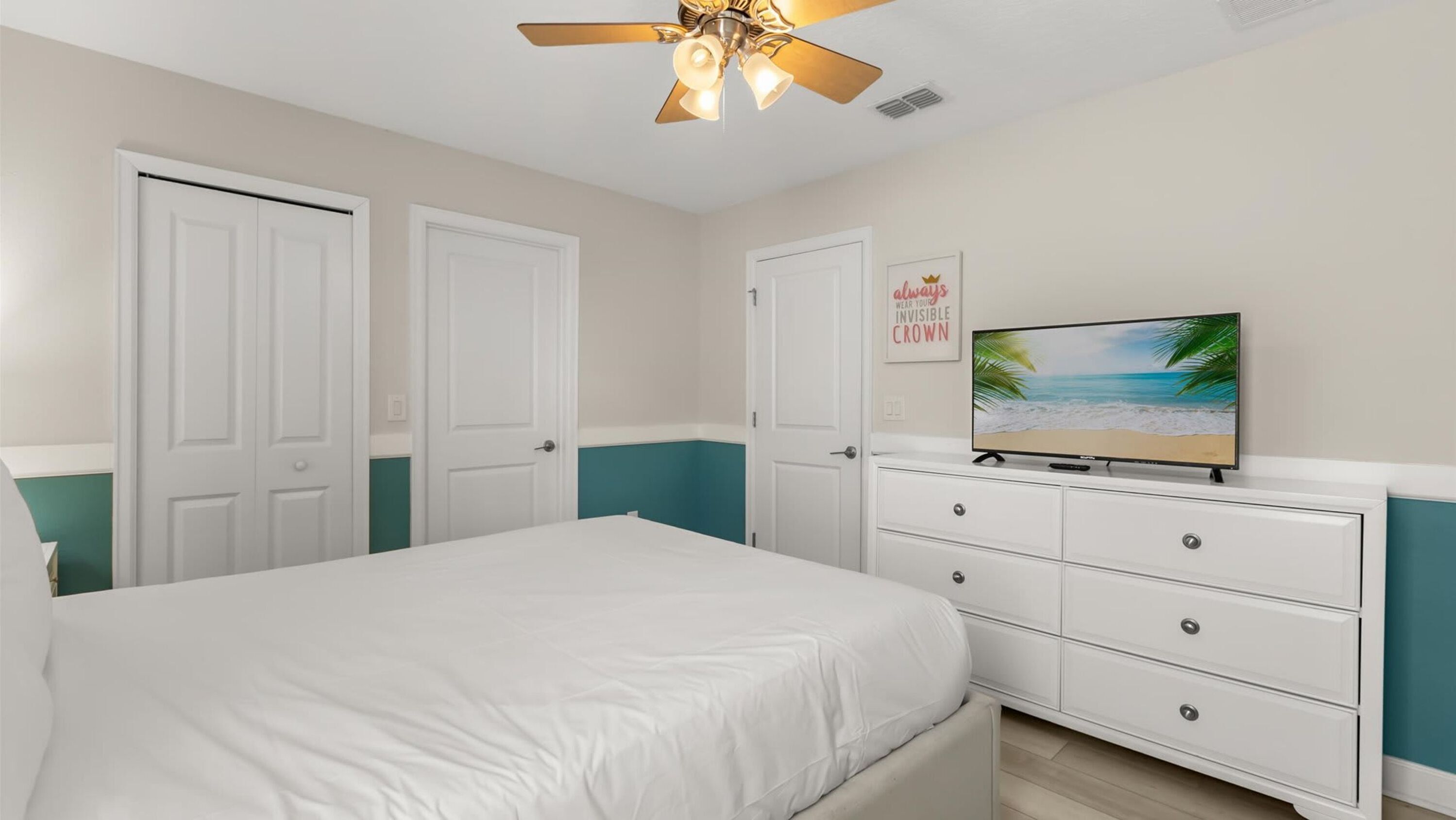 A bedroom with a bed, a dresser, a ceiling fan, and a TV displaying a beach scene.
