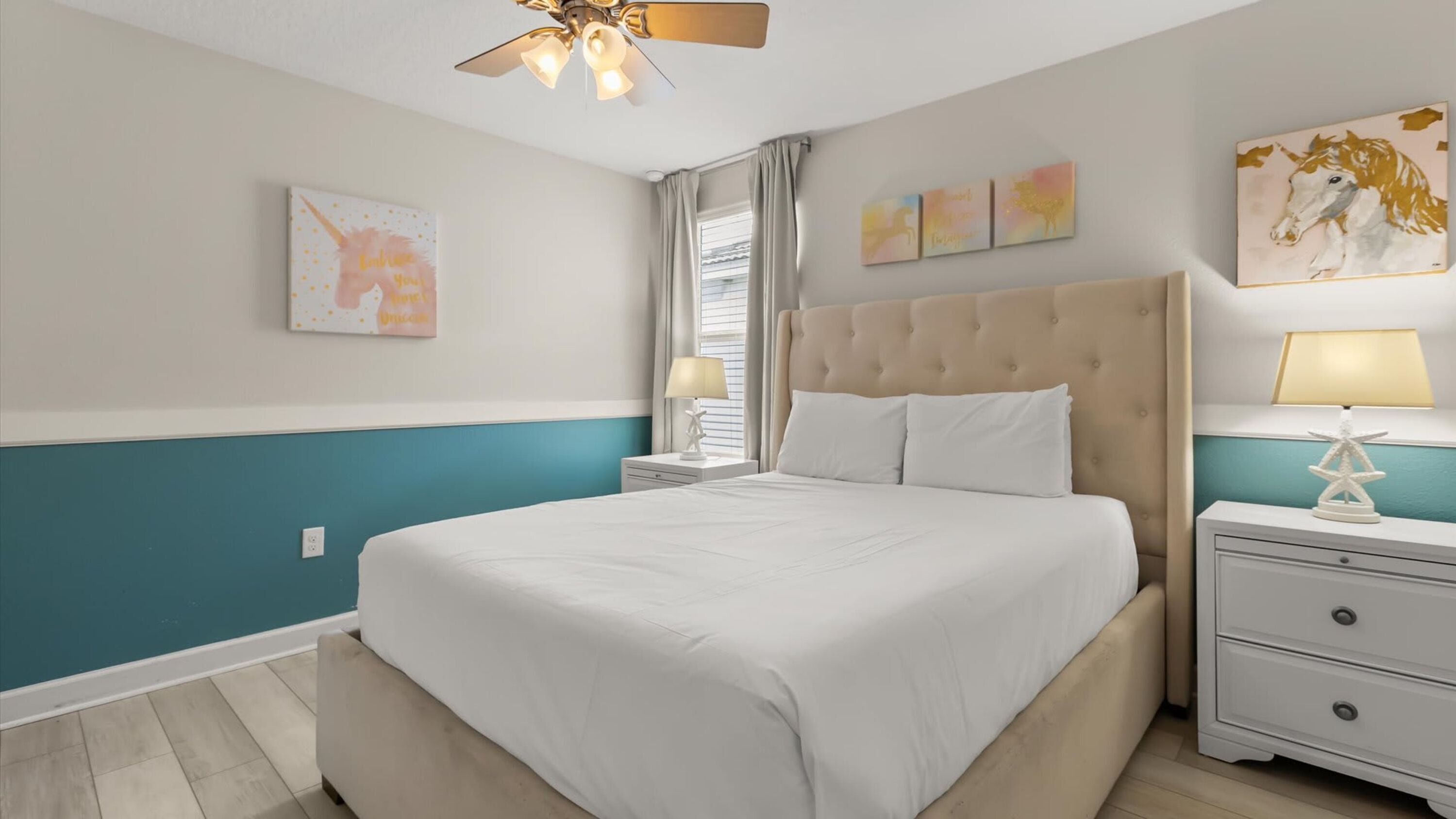A bedroom with a large bed, a ceiling fan, a nightstand with a lamp, and artwork on the walls.