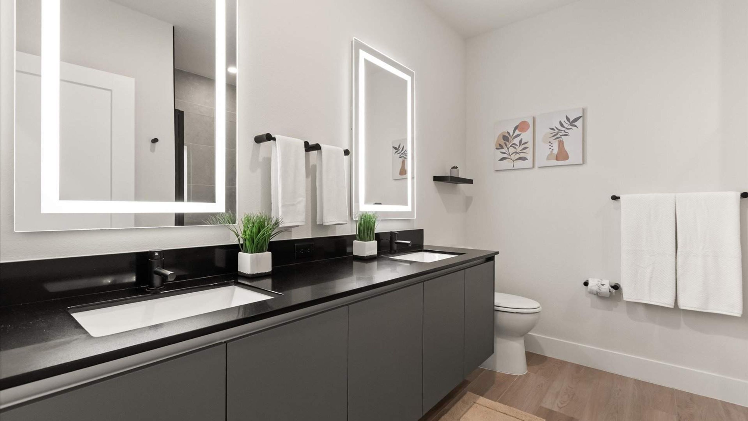 Un baño moderno con encimera negra, dos lavabos, un inodoro y un espejo grande. Hay dos plantas en macetas sobre la encimera y un perchero con toallas.