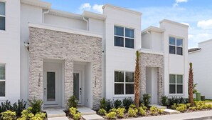 A modern two-story residential building with stone facade accents, multiple windows, and a well-maintained front yard with shrubs and palm trees. - Upstay-luxury 3BR Home w Private Pool Games Room (Clermont)