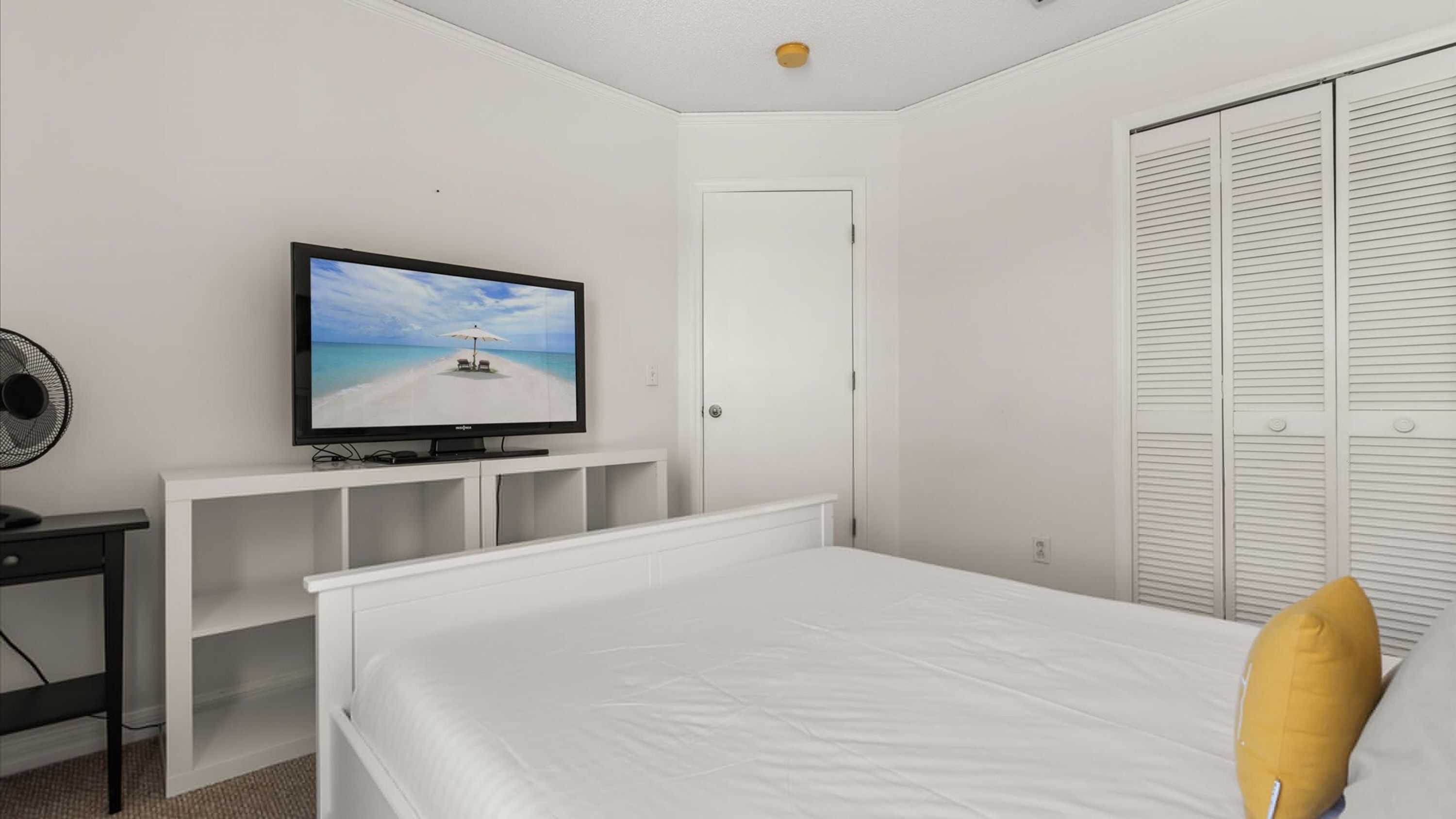 Une chambre à coucher avec un lit, un téléviseur posé sur un meuble blanc, un ventilateur et une armoire à portes à persiennes.