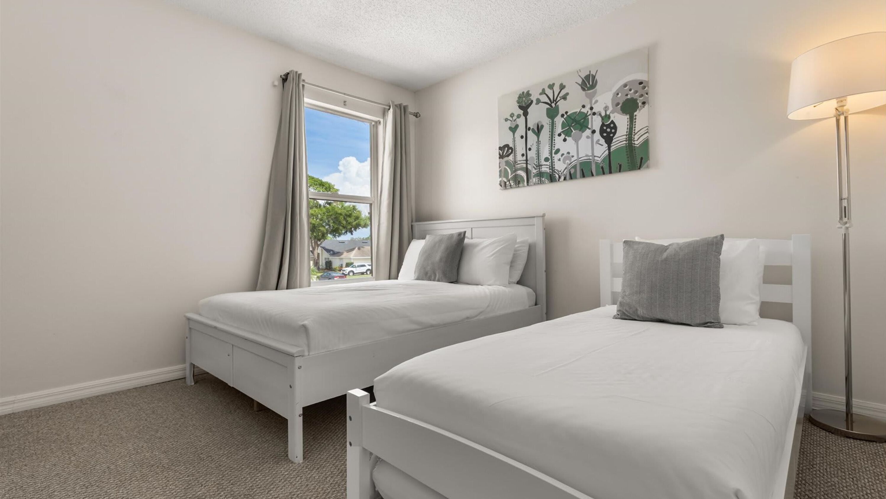 A bedroom with two single beds, a window with curtains, a painting on the wall, and a floor lamp.