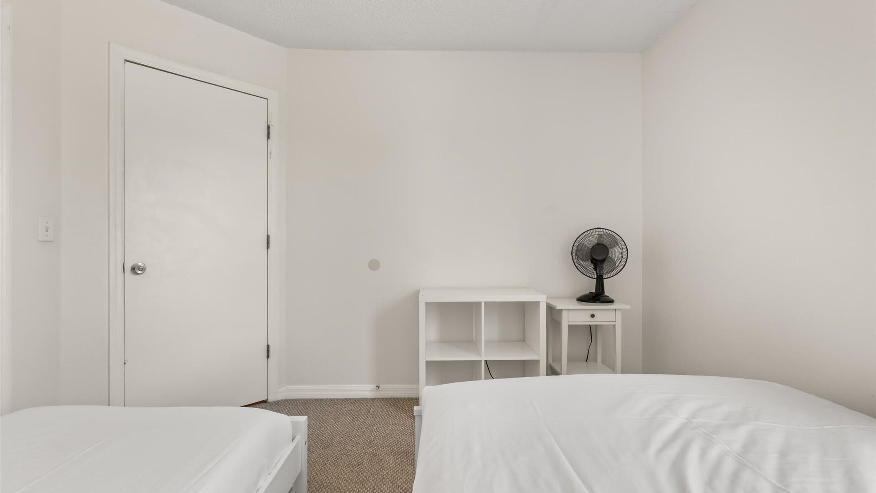 A simple, clean room with two beds, a white wardrobe, and a small desk with a fan.