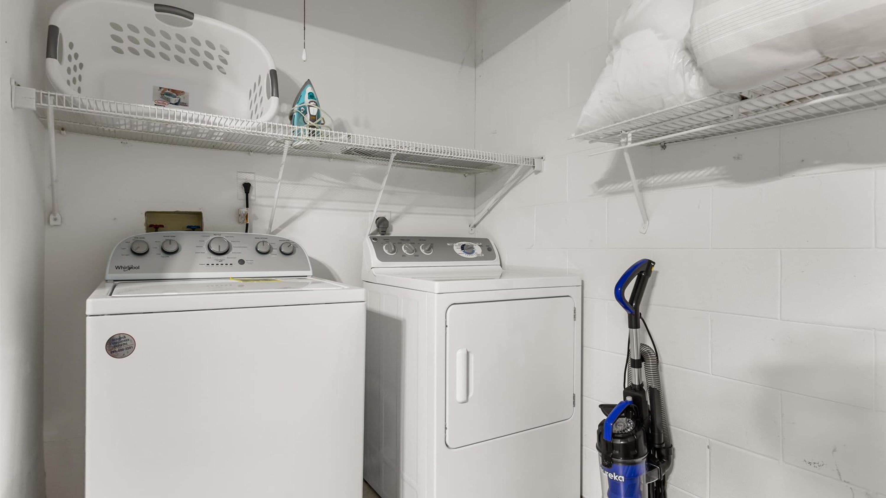 A laundry area with two washers and dryers, a shelf with a basket and a toy, and a vacuum cleaner.