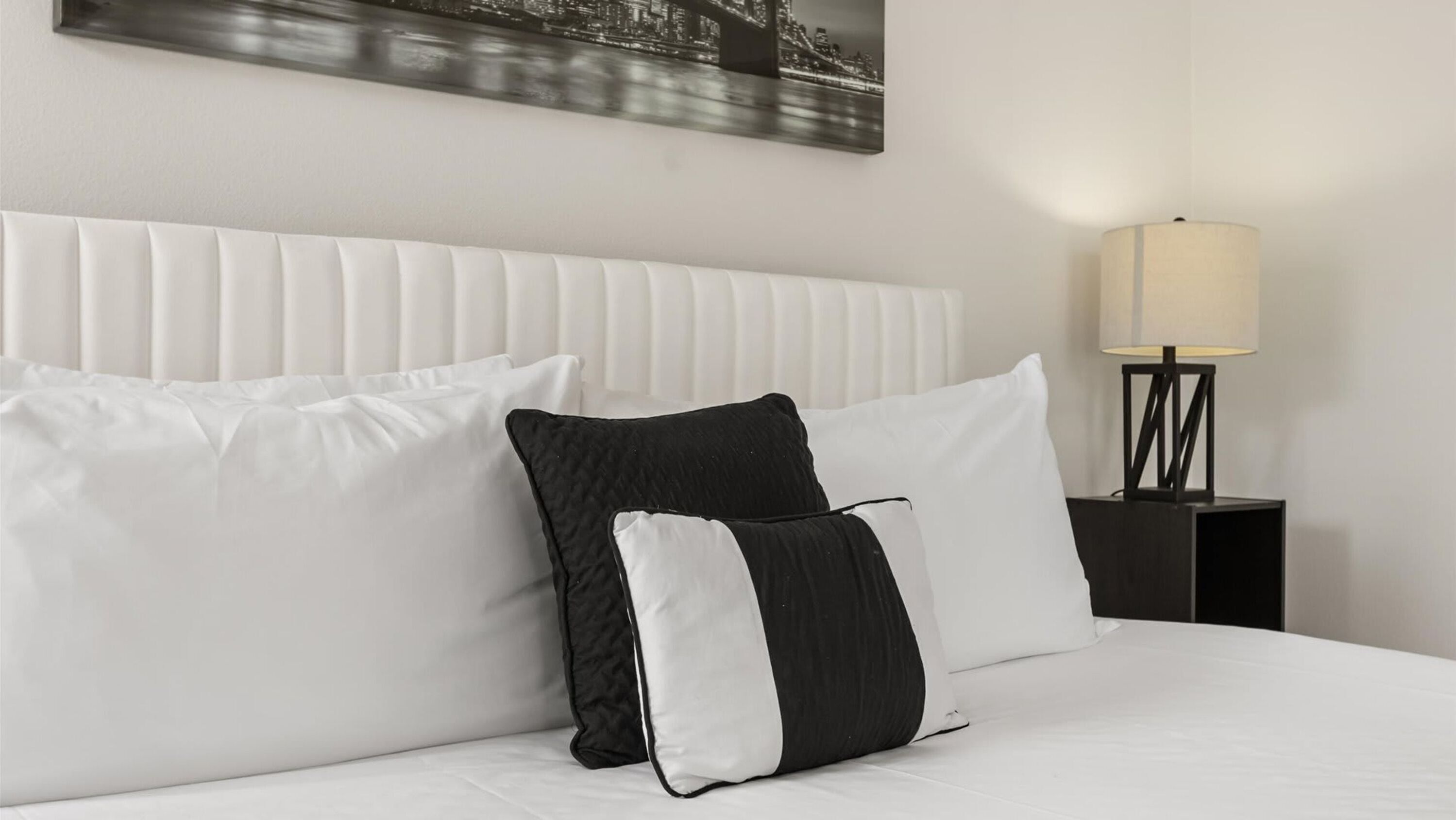 Una cama bien hecha con almohadas blancas y negras, un cabecero blanco y una mesita de noche con una lámpara.
