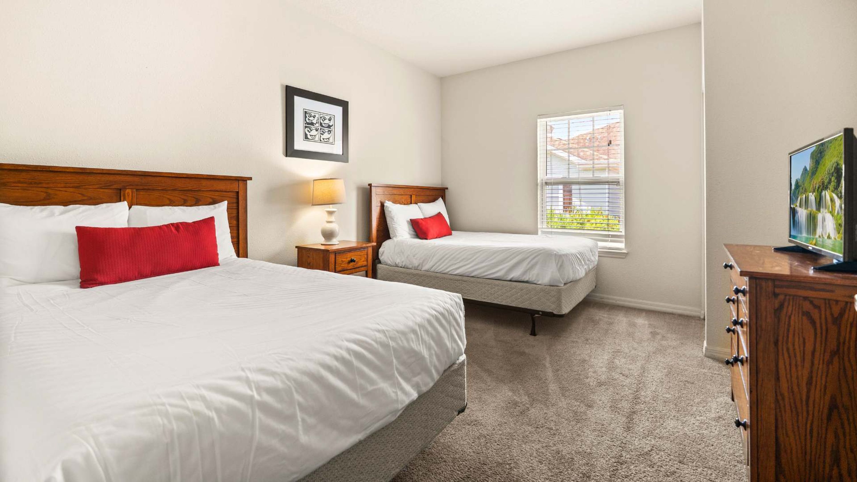 A hotel room with two beds, a nightstand with a lamp, a dresser, a window with a view, and a framed picture on the wall.