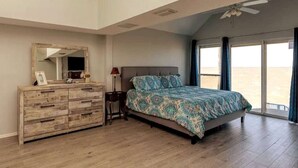 Una camera da letto con un letto grande, una cassettiera in legno, un comodino, una ventola a soffitto e vista sulla spiaggia dalla finestra.