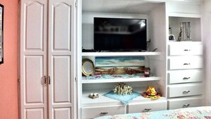 Ein Schlafzimmer mit Fernseher auf einem Schrank, einem Bett mit blumigem Überwurf und einem weißen Kleiderschrank mit mehreren Schubladen.