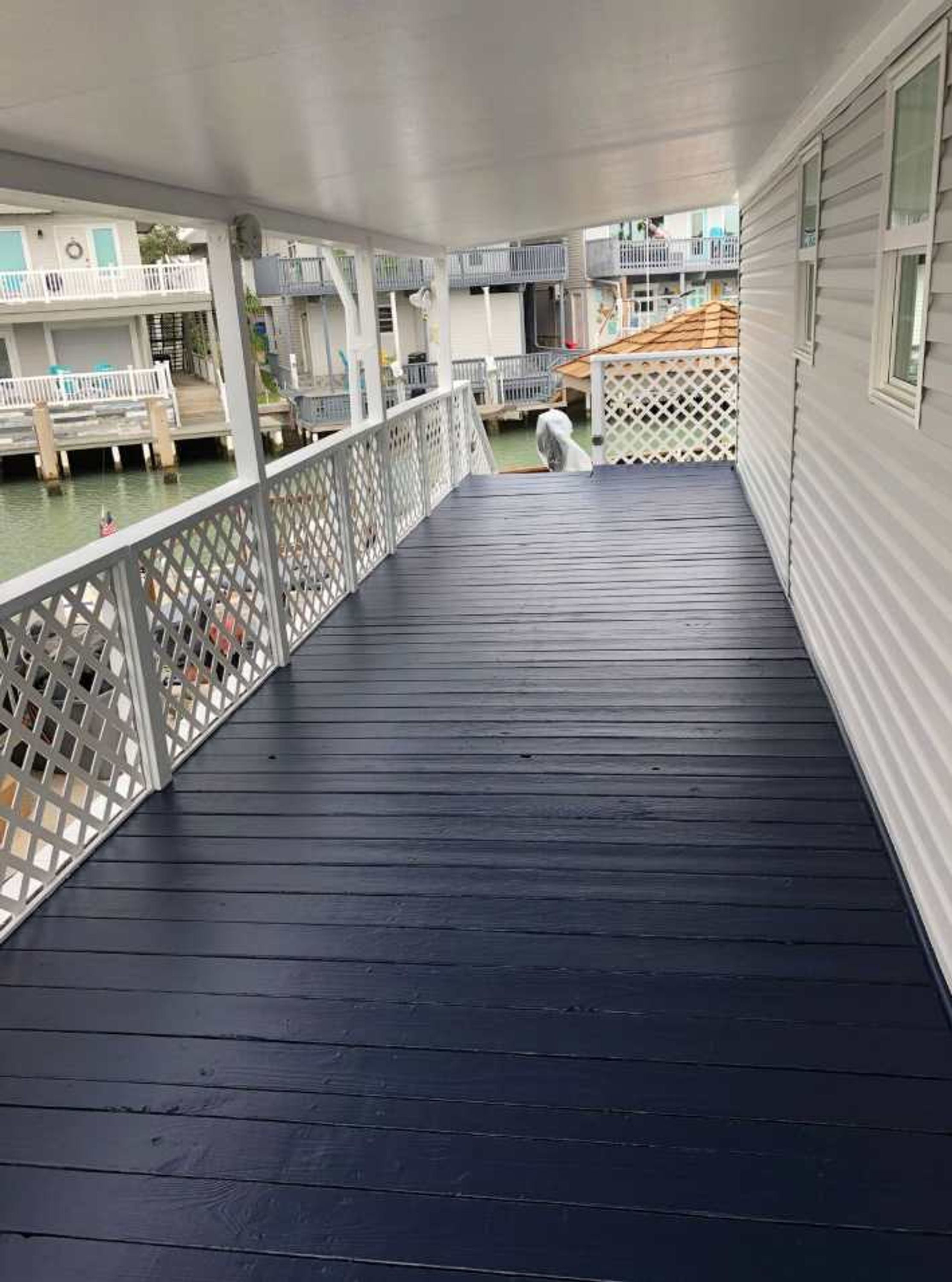 A covered outdoor deck with a wooden floor, white railings, and a view of the water and buildings in the background.