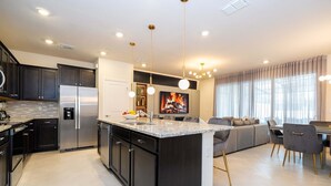 A modern kitchen with dark cabinetry, stainless steel appliances, a central island, pendant lighting, and a dining area with a gray sofa and chairs. - Upstay Family Fun 5BR Retreat w Pool Games Room (Clermont)