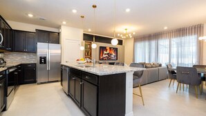 A modern kitchen with dark cabinetry, a stainless steel refrigerator, and a central island with a sink. There is a dining area with a table and chairs, and a living room with a sofa and a fireplace. - Upstay Family Fun 5BR Retreat w Pool Games Room (Clermont)