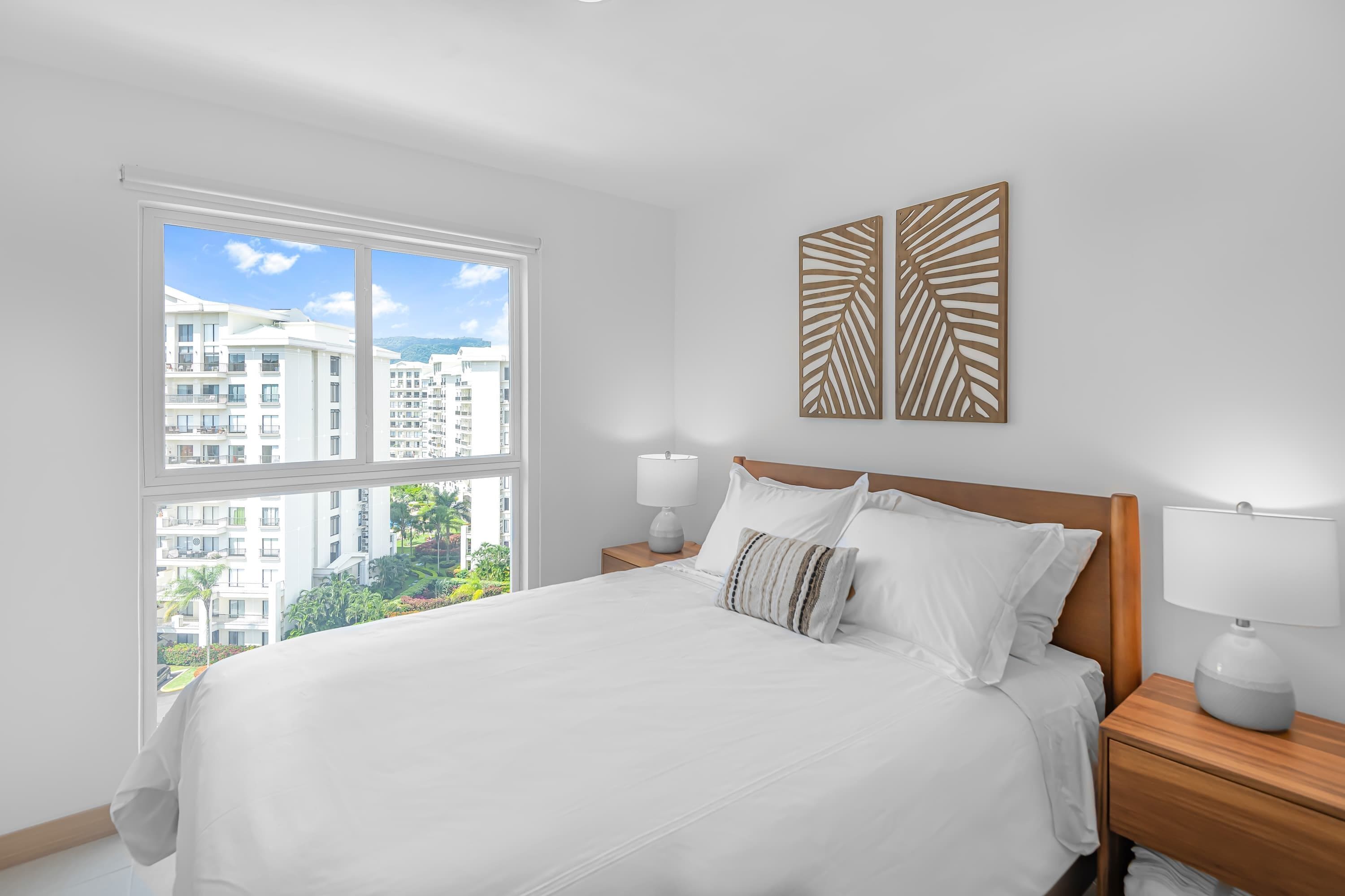 A modern hotel room with a large bed, two bedside tables with lamps, a window with a city view, and decorative wall art.