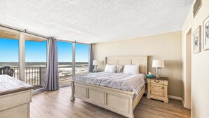Un dormitorio con una cama grande, una mesita de noche y vistas a la playa a través de una puerta corredera.