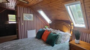Ein gemütliches Zimmer mit Holzbalkendecke, einem Bett mit gemusterten Textilien und einem Nachttisch mit Lampe und Pflanze.
