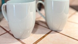 Dos tazas blancas de cerámica sobre una encimera de cocina con azulejos, con una cafetera y utensilios de cocina al fondo.
