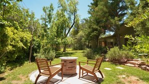 Un patio al aire libre con una mesa redonda y dos sillas de madera, rodeado de abundante follaje y árboles.