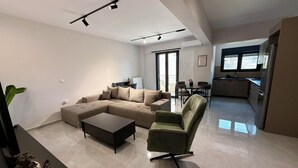 A modern living room with a sectional sofa, a coffee table, and a green armchair. There is a dining area with a table and chairs, and a kitchen in the background. - Comfort SpataAir Stay (Spata)