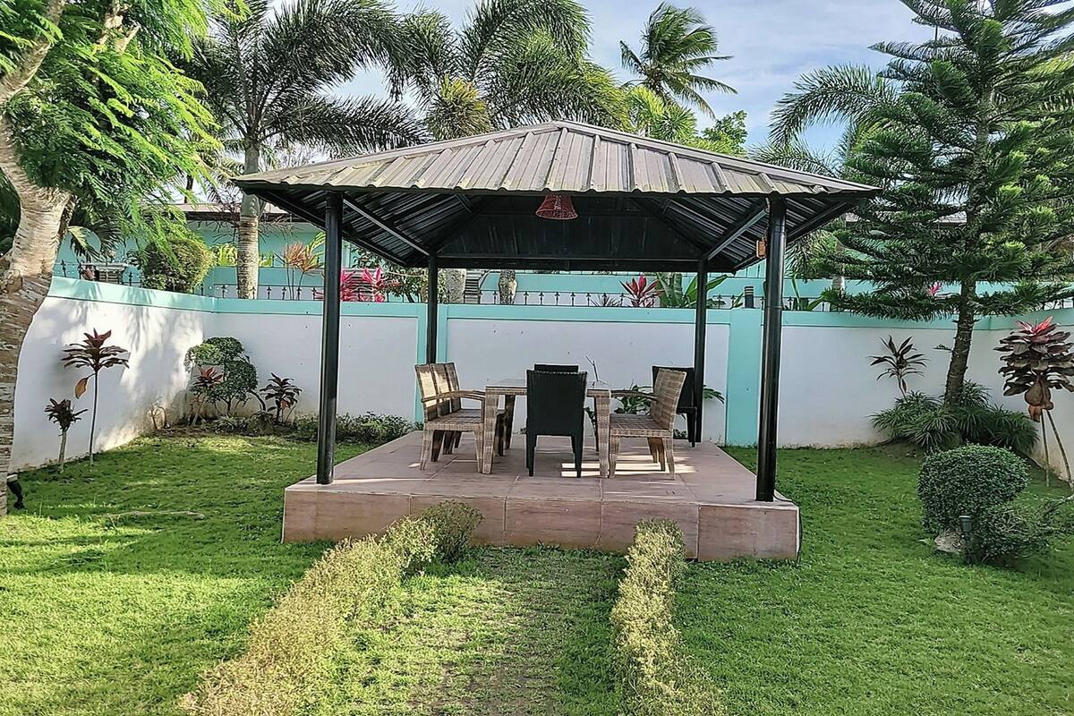 A covered outdoor seating area with a table and chairs, surrounded by a low wall and lush greenery.