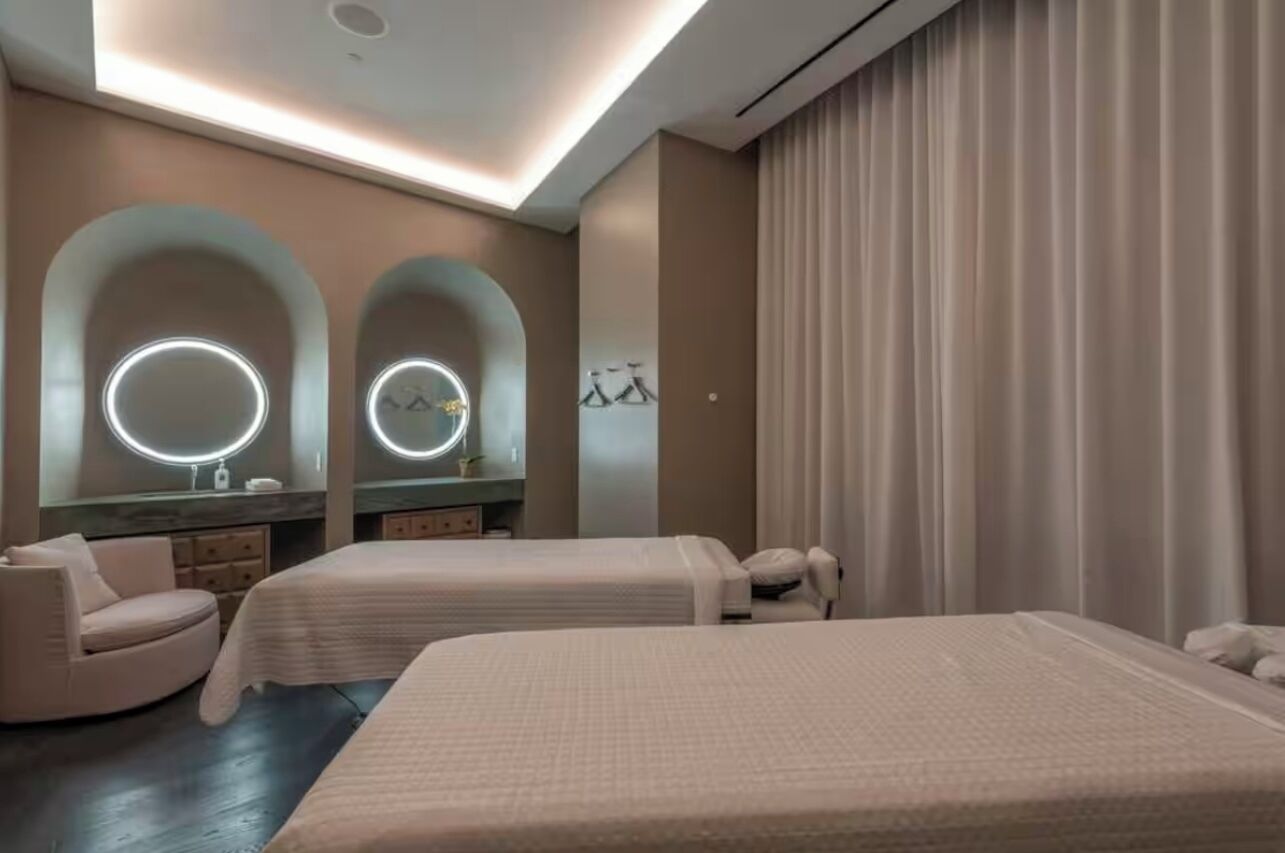Quarto de hotel moderno com duas camas, uma toucador com espelhos e uma área de estar circular.