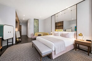 Une chambre d’hôtel moderne dotée d’un grand lit, d’une table de chevet avec une lampe, d’un coin salon avec un canapé et des fauteuils, et d’un grand miroir.