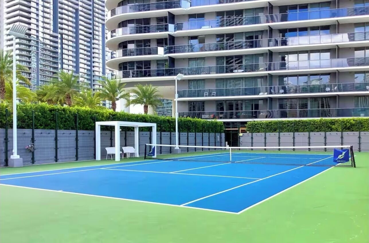 Ein Außen-Tennisplatz mit Netz, umgeben von einer niedrigen Hecke und einem Gebäude mit Balkonen.