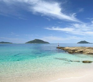Een strand met helder turquoise water, een rotsformatie en iemand die aan het vissen is.