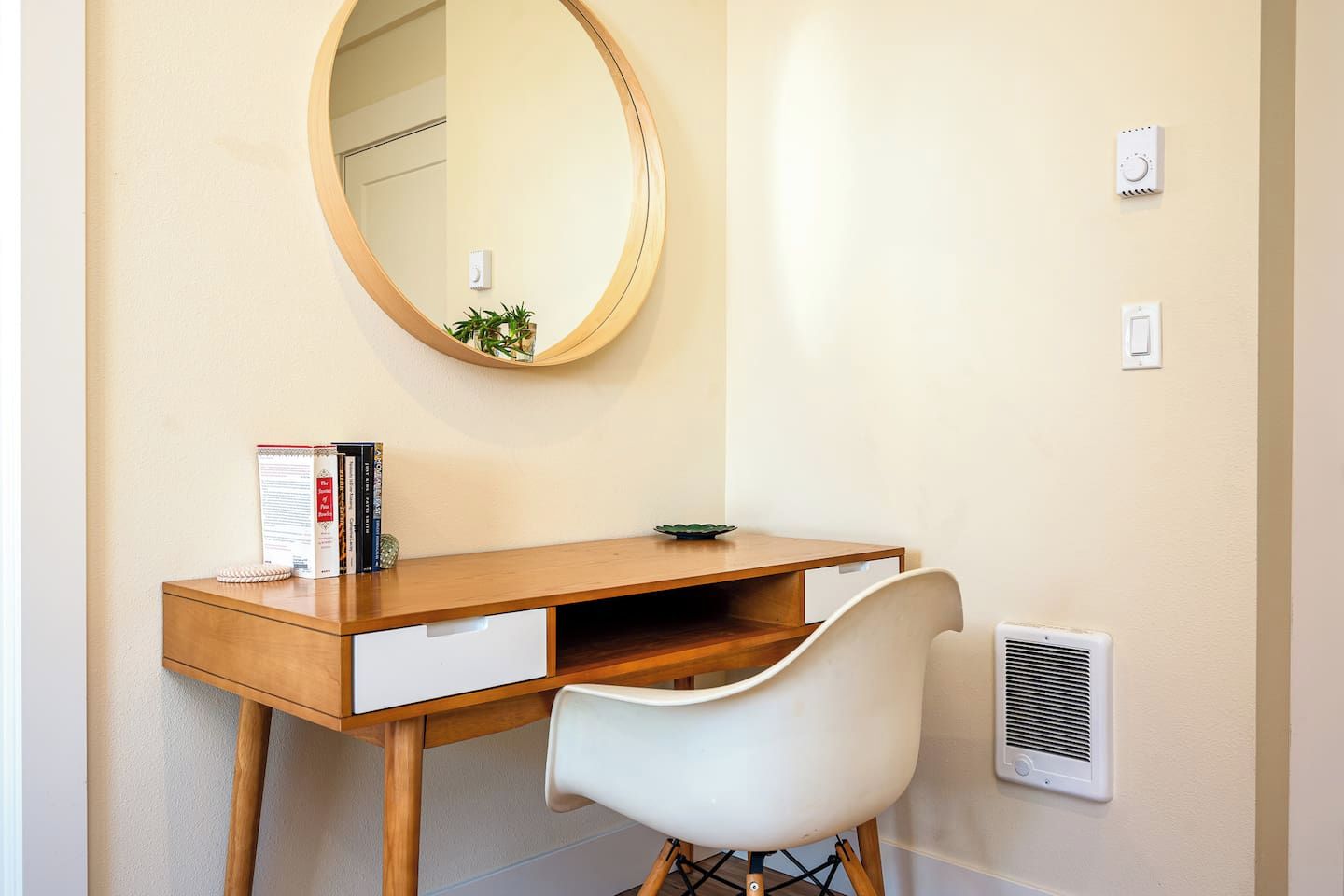 A compact bedroom with a wooden desk, a white chair, a round mirror, and a small potted plant.