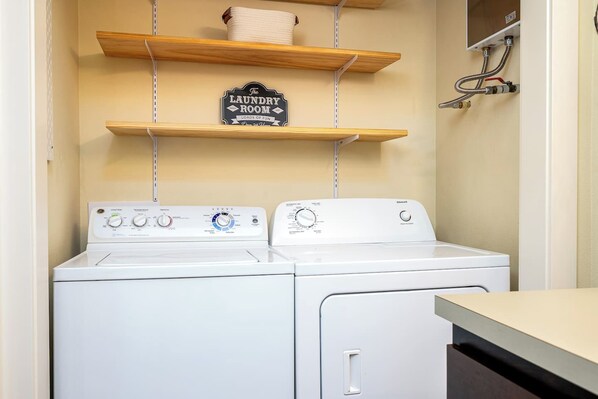 A laundry room with two white washing machines, wooden shelves, and a sign that reads 'The Laundry Room Loads of Fun.'