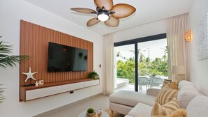 A modern living room with a flat-screen TV, wooden panel wall, ceiling fan, sofa, coffee table, and a view of greenery outside. - Luxury 2BR apartment steps from the beach (Punta Cana)