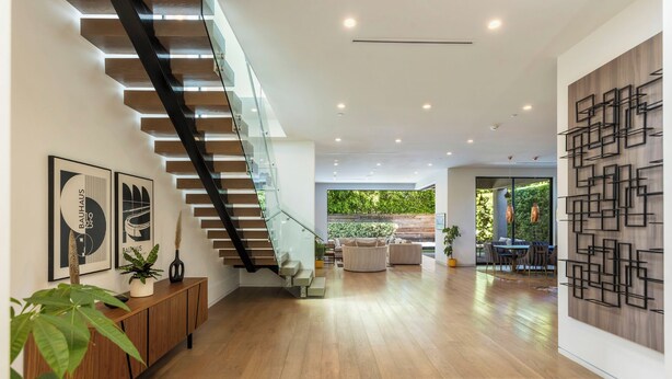 Un intérieur moderne avec un escalier en bois, un coin repas offrant une vue sur la verdure et un mur orné d’œuvres d’art géométriques.