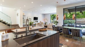 Une cuisine moderne dotée d’un îlot central, un coin repas avec une grande table et des chaises, un salon avec un canapé et une télévision, et un escalier avec des rampes en verre.