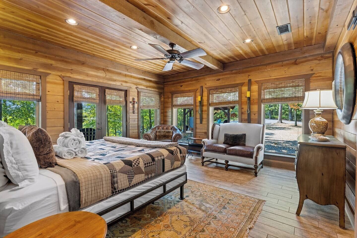 A log cabin bedroom with a bed, two chairs, a desk, and a view of the outdoors.