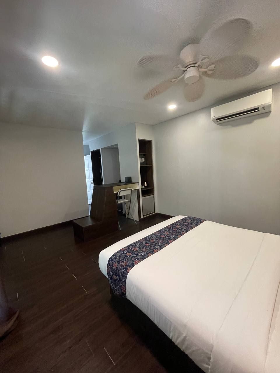 A hotel room with a bed, a desk, a chair, a ceiling fan, and an air conditioning unit.