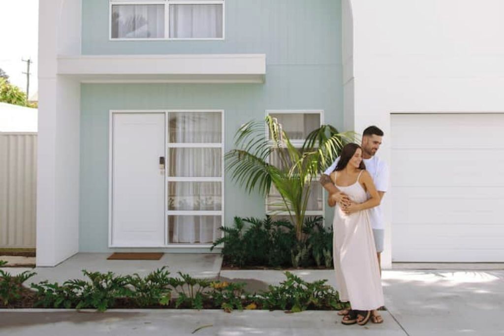 A two-story house with a light blue exterior, white garage door, and a small garden with plants and a palm tree.