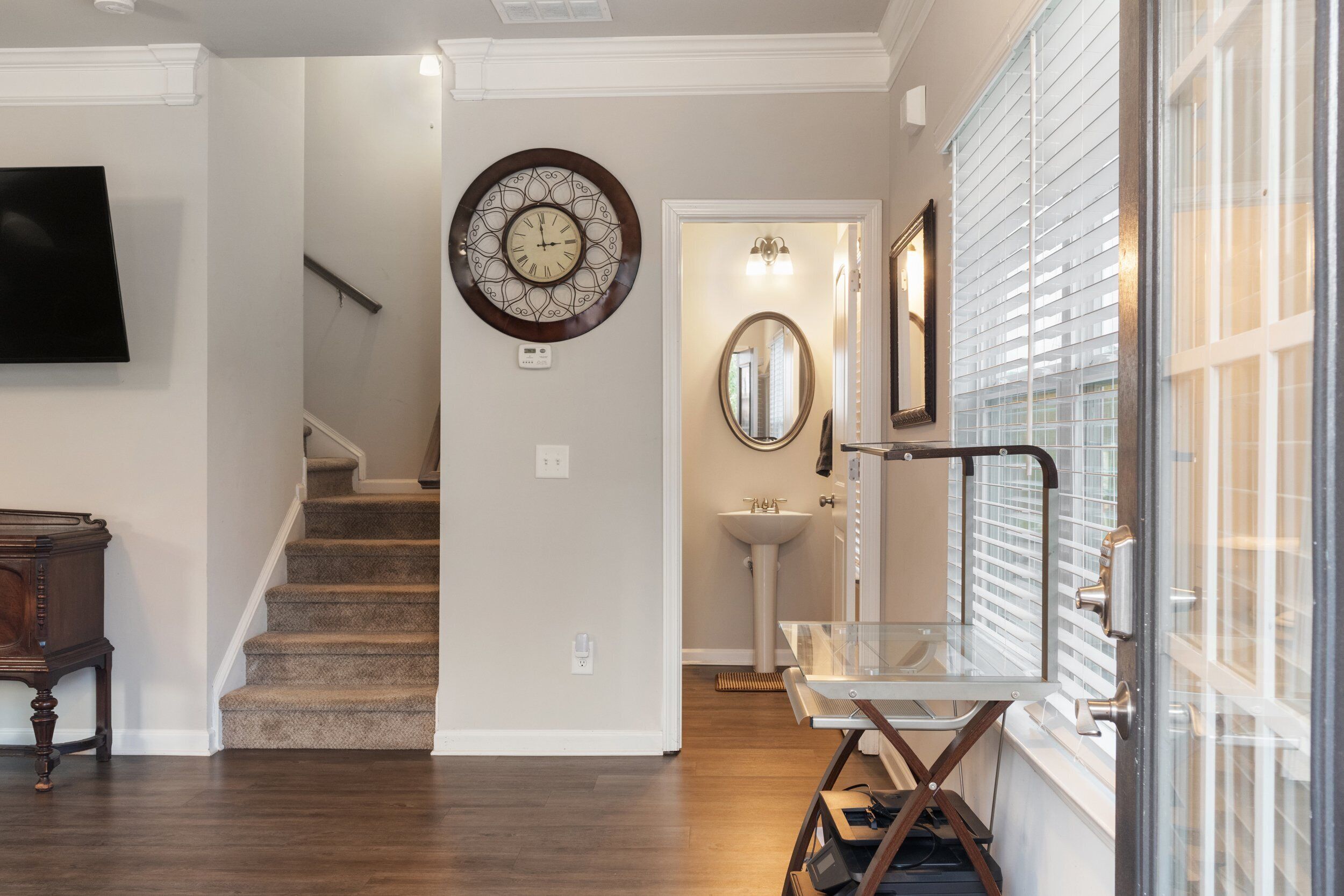 A hallway with a staircase, a wall-mounted clock, a mirror, and a glass door leading to a bathroom.