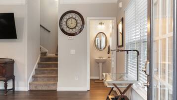 A hallway with a staircase, a wall-mounted clock, a mirror, and a glass door leading to a bathroom.