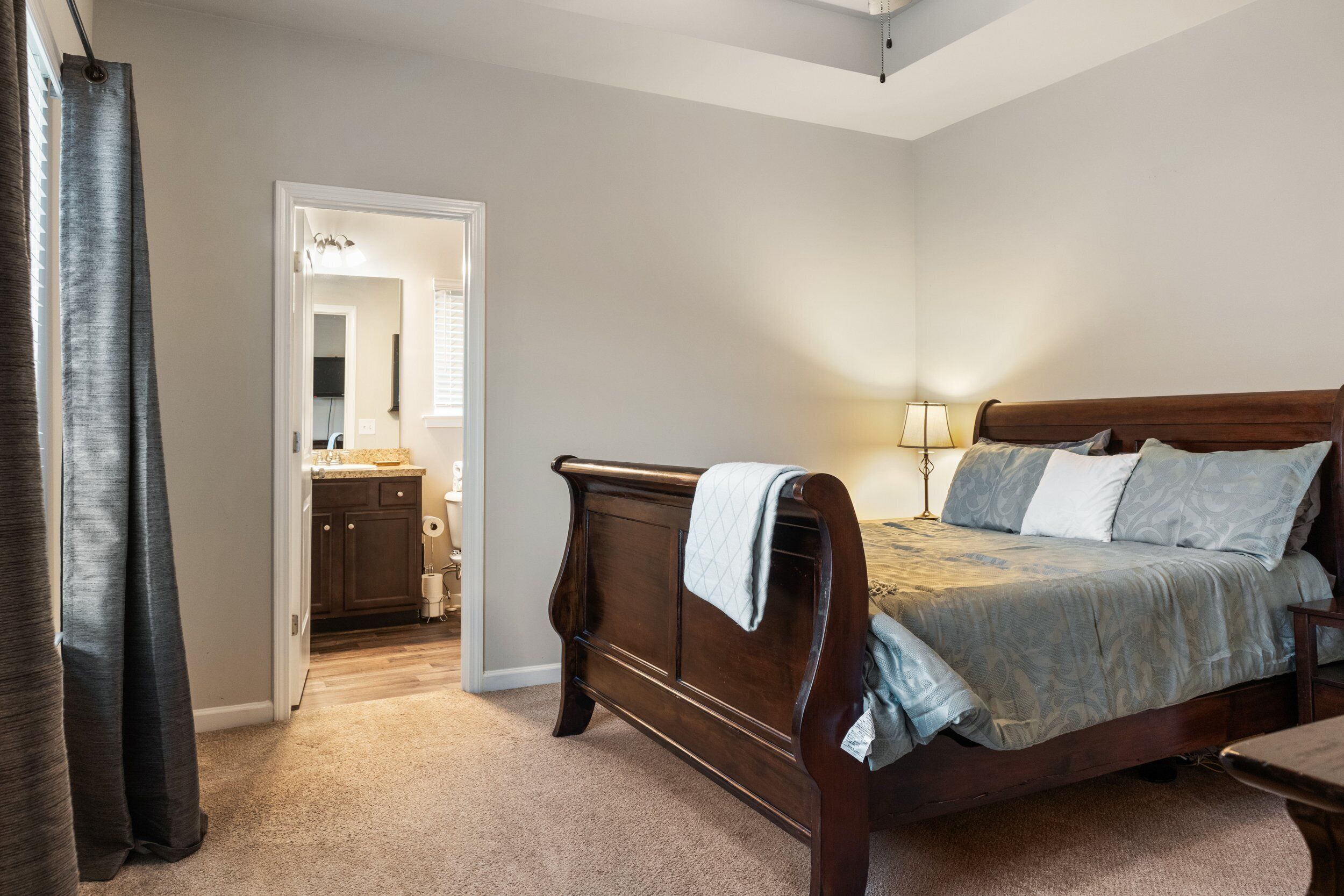 A bedroom with a wooden bed, a nightstand, and a lamp. There is a door leading to a bathroom.