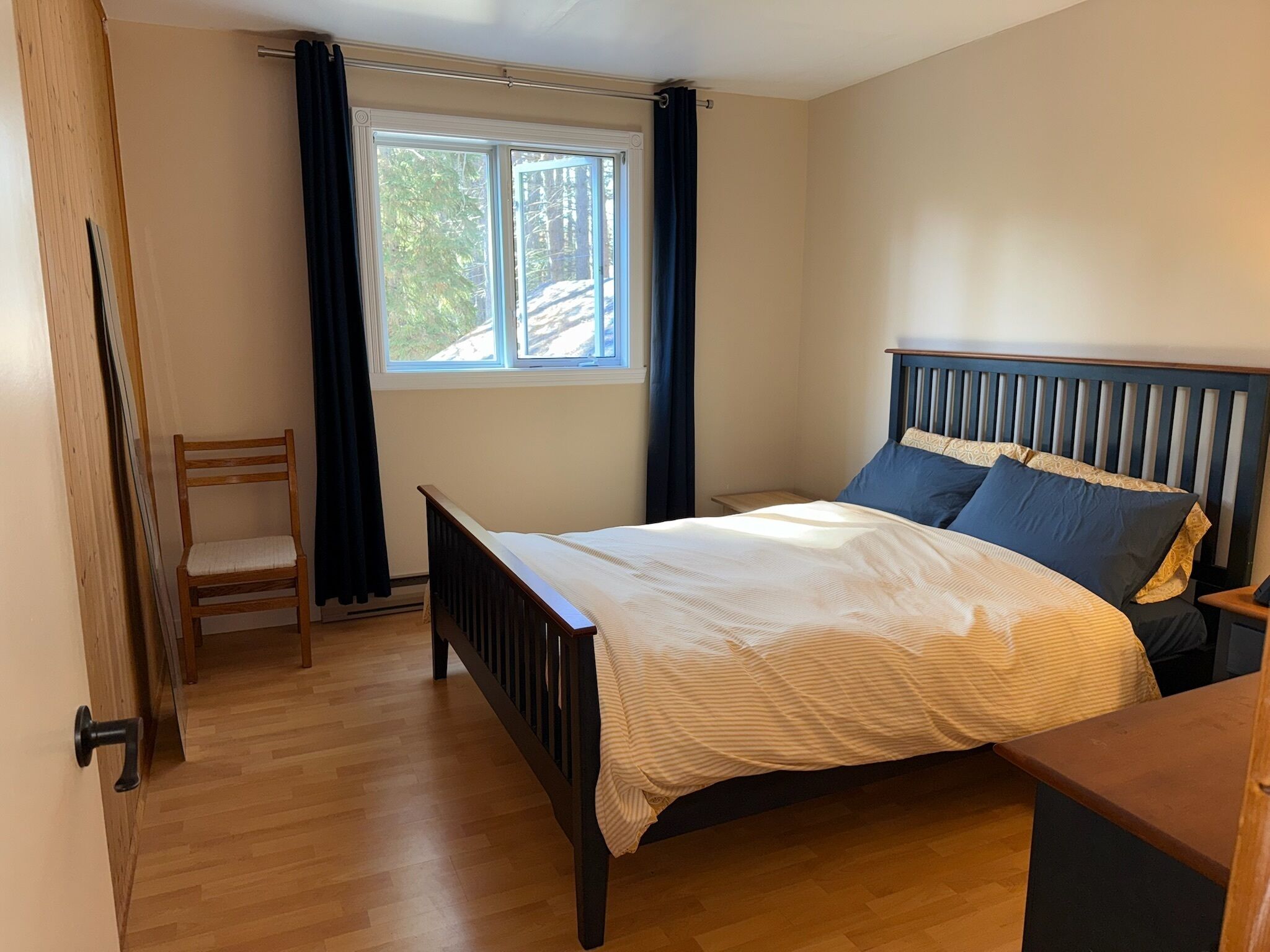 Un dormitorio con una cama de madera, una silla, un ventanal con cortinas y vistas a árboles.