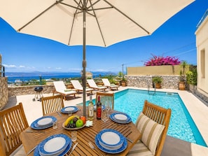 Un espace repas aménagé au bord de la piscine, avec une table dressée pour quatre personnes, un barbecue et une vue sur la mer.