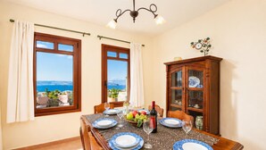 Une salle à manger avec une table dressée pour quatre personnes, deux fenêtres avec des rideaux, un lustre et un buffet en bois doté de portes en verre.