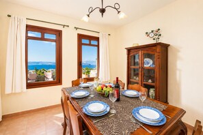 Une salle à manger avec une table dressée pour quatre personnes, deux fenêtres avec des rideaux, un lustre et un buffet en bois doté de portes en verre.