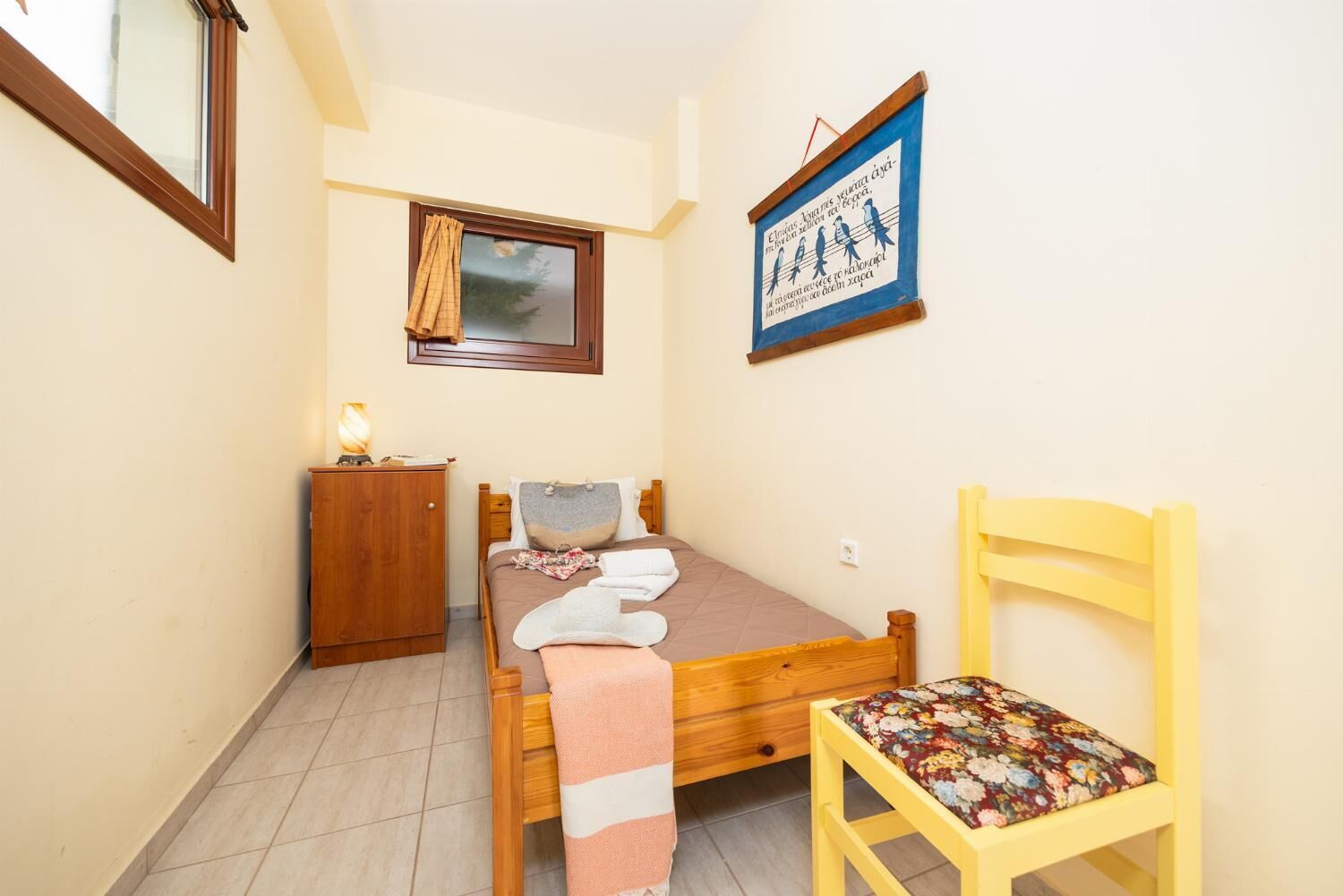 A small, cozy bedroom with a single bed, a wooden nightstand, a chair, and a window with curtains.