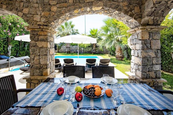 An outdoor dining area with stone pillars, a pool, and chairs.