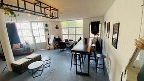 A spacious room with a long table, multiple chairs, a sofa, and a small table with a plant. There are large windows allowing natural light, and a ceiling with exposed beams and hanging lights. - Music Guest House (Mexico City)