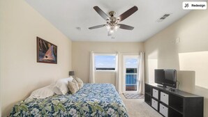 A bedroom with a bed, a ceiling fan, a television on a stand, and a balcony with a view.