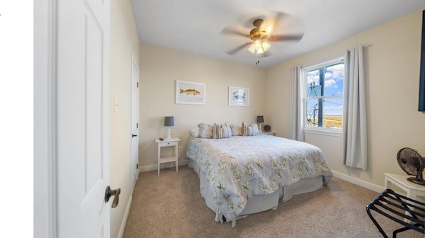 A bedroom with a bed, a ceiling fan, a window with curtains, and a small table with a lamp.